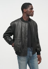 Μαύρο δερμάτινο μπουφάν τύπου bomber με υφή, ριμπ μανσέτες και διπλό φερμουάρ μπροστά, σε συνδυασμό με μαύρο T-shirt και τζιν παντελόνι.