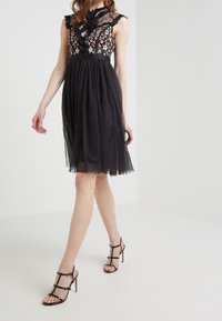 Robe noire avec broderie florale, jupe en tulle transparent et col en dentelle à volants. Associée à des sandales à talons hauts noires à lanières.