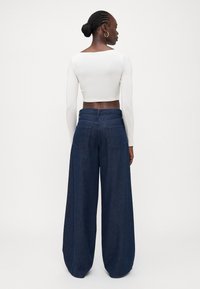 Top corto de manga larga blanco combinado con pantalones de mezclilla de pierna ancha en azul oscuro que cuentan con bolsillos traseros y una cinturilla alta. Sin patrones notables.