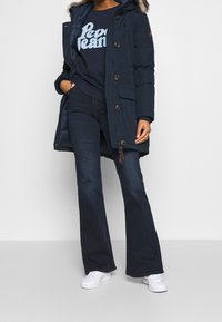 Kvinna som bär en mörkblå parka med pälsmössa, marinblå sweatshirt med "Pepe Jeans"-logotyp, mörka utställda jeans och vita sneakers, stående med händerna i fickorna.