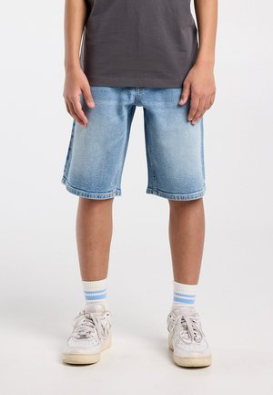 Child wearing light wash denim shorts, white sneakers, and white socks with blue stripes, standing against a plain background.