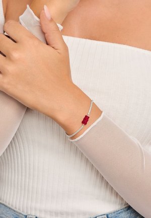 Bracciale d'argento con gemme trasparenti e un charm a forma di orsetto gommoso rosso, posizionato su uno sfondo di tessuto chiaro con texture rigata.