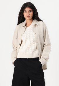 Beige skjorte med knapper og let tekstur, båret over en cremefarvet kabelstrikket sweater og sorte bukser, med en brystlomme og minimal detaljering.