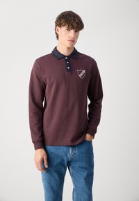 Cămașă polo cu mânecă lungă de culoare vișinie, cu guler bleumarin, placă cu trei nasturi și logo brodat în formă de scut pe pieptul stâng. Asortată cu blugi albaștri.