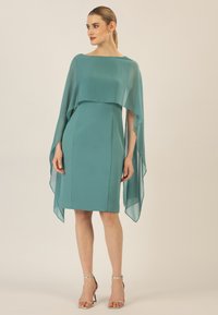 Türkises Kleid aus glattem Stoff, mit einer mehrlagigen, transparenten Überlegung. Knielang mit einer taillierten Silhouette und minimalem Zubehör.