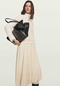 Černá kabelka s embosovaným krokodýlím vzorem a dvěma uchy. Modelka má na sobě bílý svetr s copánkovým vzorem a splývavou krémovou sukni. Jednoduchý, strukturovaný design.