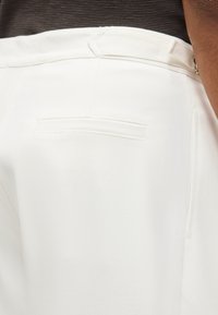 Κοντινή λήψη της πίσω τσέπης λευκού παντελονιού με θηλιές για ζώνη, που φοριέται με σκούρο καφέ τοπ.