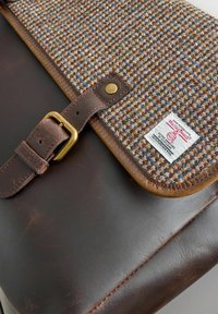 Brown leather bag with a woven wool flap, featuring a brass buckle and tag, and a textured surface with a multi-colored pattern.