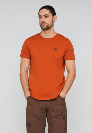 Homme portant un t-shirt orange à manches courtes et un pantalon cargo marron, les mains dans les poches, debout devant un fond clair uni.