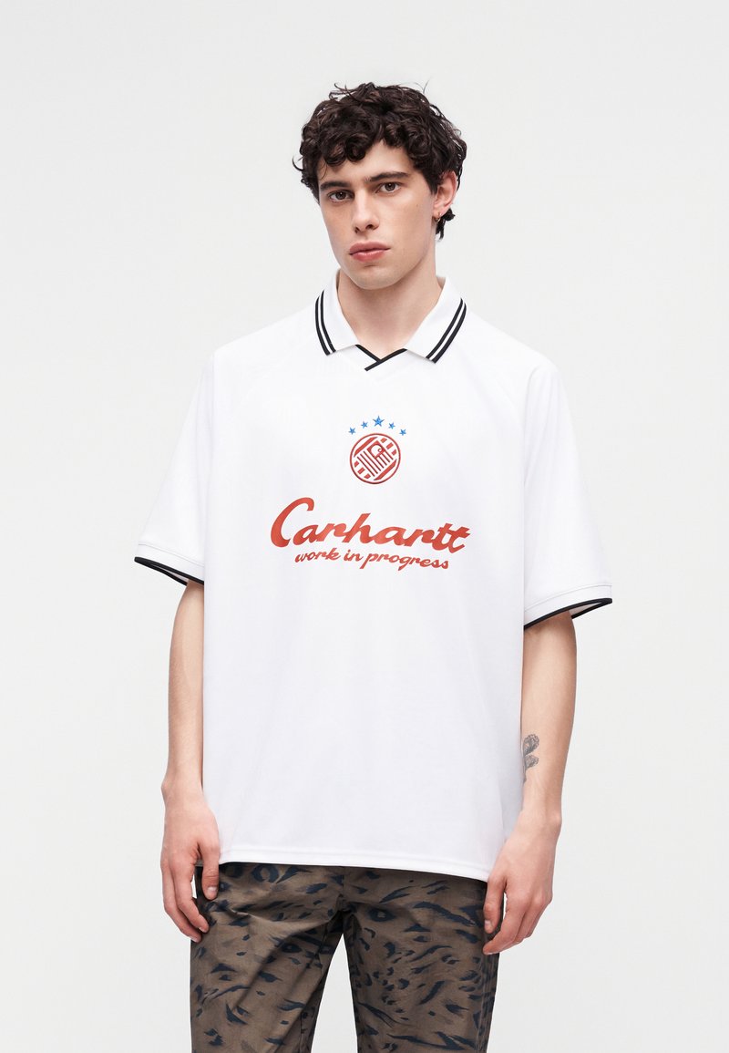 Junger Mann mit lockigem, dunklem Haar, der ein weißes Carhartt-Poloshirt mit schwarzem Rand und gemusterte braune Hose vor einem einfarbigen Hintergrund trägt.
