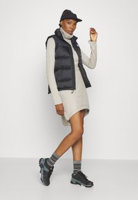 Gilet rembourré noir porté sur une robe beige clair, associé à des chaussettes rayées grises et des chaussures de sport noires. Design simple et style décontracté.