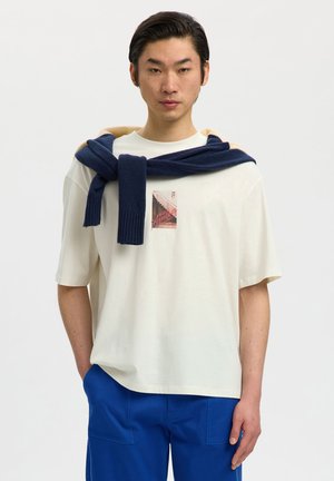 Młody mężczyzna stoi z rękami w kieszeniach niebieskich spodni, ubrany w kremowy t-shirt z grafiką i granatowy sweter zarzucony na ramiona.