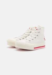 Biele tenisky s vysokým strihom vyrobené z plátna, s kovovými očkami, gumovou podrážkou a červenými akcentmi na päte a strednej podrážke.