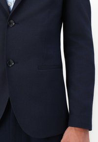 Μπλέιζερ σε ναυτικό χρώμα από λείο ύφασμα με κλείσιμο δύο κουμπιών, περασμένα πέτα και μία θήκη στην τσέπη του στήθους. Το μοντέλο δείχνει τις λεπτομέρειες των μανικιών.