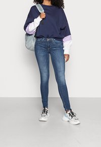 Sudadera oversized de color navy con mangas moradas y blancas, combinada con jeans skinny azules y zapatillas blancas con acentos en negro y azul.