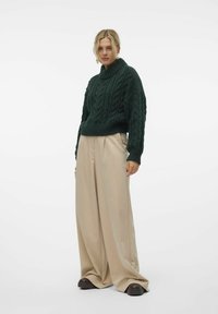 Donkergroene kabelgebreide coltrui met lange mouwen, gecombineerd met brede beige broek en zwarte chunky laarzen. Glad textuur.