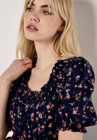 Rückenfreies Kleid aus marineblauem Stoff mit Puffärmeln und gerafftem Ausschnitt. Mit kleinen rosa und grünen Blumenmustern durchzogen.