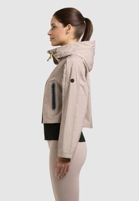 Kvinde iført beige hættetrøje med lynlås og sort top, vist i profil mod en ensfarvet baggrund.