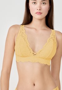 Bralette giallo in pizzo con design a scollo a V, spalline regolabili e bordi scallopati. Presenta un dettaglio incrociato anteriori. Texture morbida.