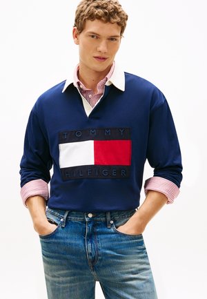 Giovane uomo con capelli ricci che indossa una felpa Tommy Hilfiger blu navy sopra una camicia a righe rosse e bianche e jeans blu, con le mani nelle tasche.