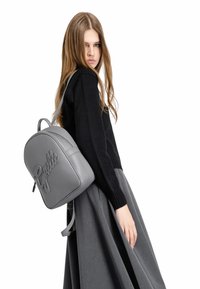 Zaino in pelle grigia con forma arrotondata, logo in rilievo, spallacci regolabili e texture liscia. Indossato su una spalla sopra un maglione nero.