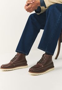 Ruskeat nahkaiset nilkkurit, joissa valkoiset kumipohjat, pyöreät nauhat ja kontrastitikkaus, yhdistetty sinisiin farkkuihin.