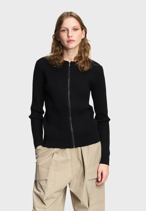 Young woman with shoulder-length wavy hair wearing a black ribbed zip-up sweater and beige cargo pants against a plain background.