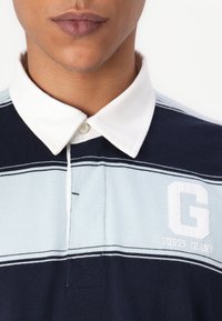 Polo tričko s námorníckym a svetlomodrým pruhom, s bielym golierom a vyšitým bielym logom "G" značky Guess Jeans. Mäkko textúrovaná tkanina.