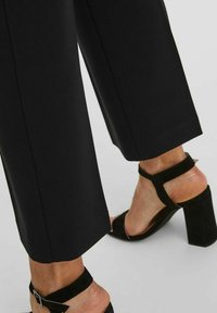 Los pantalones negros ajustados presentan un corte recto y una textura suave, combinados con zapatos de tacón alto negros con correas para el tobillo.