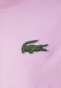 T-shirt rosa in cotone con scollatura rotonda. Presenta un logo di un coccodrillo ricamato in verde con accenti rossi nella parte sinistra del petto.