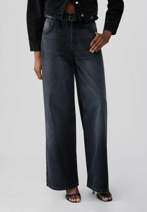 Persona che indossa jeans neri a vita alta e gamba larga, cintura nera, giacca corta nera e tacchi neri aperti sulle punte su uno sfondo chiaro uniforme.