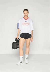 Λευκή φαρδιά μπλούζα Adidas με κόκκινες και μπλε λεπτομέρειες, μαύρο σορτς, λευκές κάλτσες και λευκά αθλητικά παπούτσια, κρατώντας μια μαύρη τσάντα γυμναστηρίου.