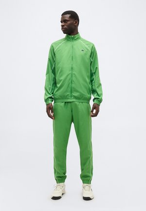 Άνδρας στέκεται κοιτώντας προς τα αριστερά, φορώντας πράσινη φόρμα με φερμουάρ και λευκή επένδυση, καθώς και λευκά αθλητικά παπούτσια, μπροστά σε ένα απλό ανοιχτό φόντο.