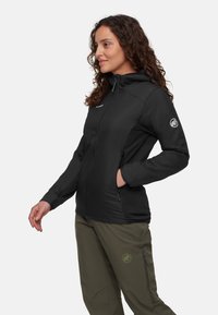 Kvinna med lockigt hår som bär en svart zip-up hoodie och olivgröna byxor, står med en hand i fickan och ett svagt leende.