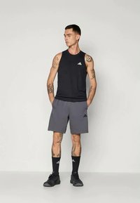 Melnas sporta bez piedurknēm un pelēki šorti ar mazu baltu logo. Modelis valkā melnas zeķes un apavus, uz rokām redzami tetovējumi.