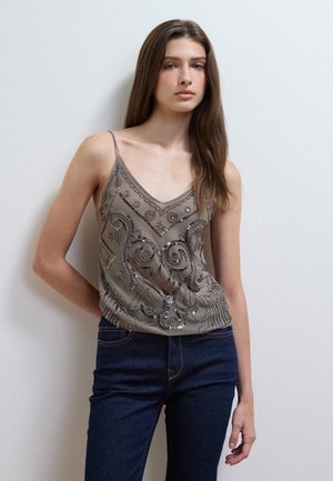 Giovane donna con capelli lunghi e castani, che indossa una canotta taupe con dettagli di perline e jeans blu scuro, in piedi contro una parete bianca semplice.