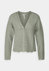 Napapaita cardigan vaaleassa vihreässä. Ominaisuuksina teksturoitu vaijeri neule, aallotettu helma ja pitkät hihat. Valmistettu pehmeästä, joustavasta materiaalista.