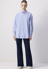 Giovane donna che indossa una camicia a righe verticali blu e bianche con bottoni, pantaloni svasati scuri e sneakers bianche, in piedi davanti a uno sfondo semplice.