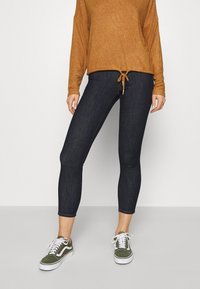 Leggings de mezclilla oscura con forma ajustada, combinadas con una camisa de manga larga marrón con detalle de cordón y zapatillas verde oliva.