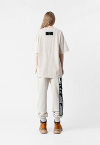 Camiseta de gran tamaño color beige con mangas cortas, que presenta una etiqueta en la parte posterior. Combinada con pantalones gris claro que tienen acentos gráficos negros y botas color tostado.