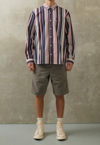 Man in een langemouwen donkerblauw, wit en rood gestreept overhemd, grijze knielange shorts, witte sokken en beige hoge sneakers.