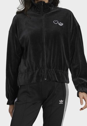 Vrouw draagt een zwart velours jack met rits, voorzien van een hart- en Adidas-logo, gecombineerd met zwarte Adidas trainingsbroek met witte strepen.