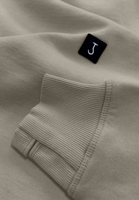 Beiger Sweatshirt mit gerippten Bündchen und einem kleinen schwarzen Etikett mit einem weißen "J". Weiche Textur, genähter Saum und detaillierte Nähte.