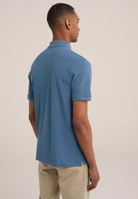 Polo majica iz mehkega modrega bombaža, kratki rokavi, zloženi manšeti, stranski razporki na robu in klasičen ovratnik. Gladka tekstura in prilegajoča zasnova.