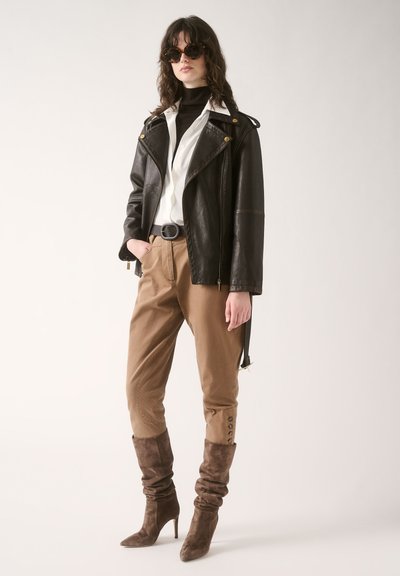 Black leather biker jacket with gold accents, white shirt, tan tapered trousers, and slouchy brown suede high-heeled boots.