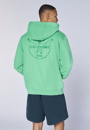 Mann trägt einen grünen Hoodie mit einem Taucher-Logo und dem Schriftzug "Chiemsee" auf dem Rücken, kombiniert mit dunkelblauen Shorts, Rücken zugewandt.
