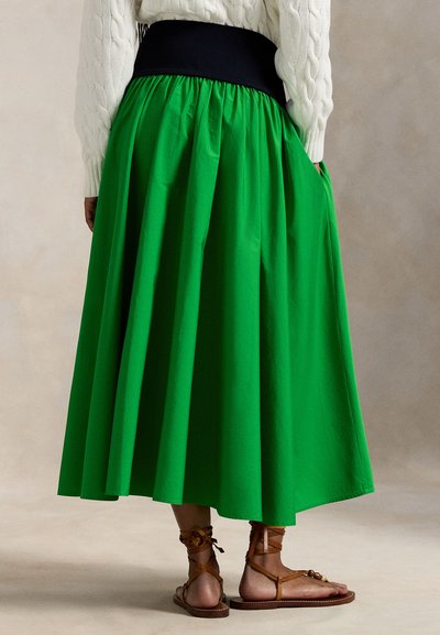 Πράσινη, ανοιχτή φούστα μήκους μέχρι τον αστράγαλο με συγκεντρωμένη ζώνη στη μέση και απαλό ύφασμα. Συνδυάζεται με καφέ σανδάλια με λουριά που αφήνουν εμφανείς λεπτομέρειες στους αστραγάλους.