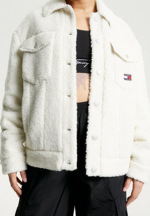 Personne portant une veste blanche duveteuse Tommy Jeans ouverte sur un haut court noir et un pantalon noir, montrant un tatouage sur le ventre.