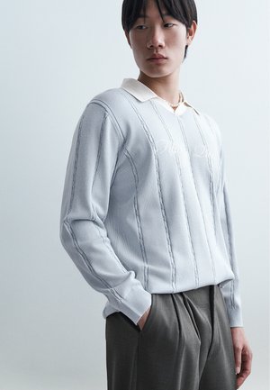 Jeune homme aux cheveux noirs portant un pull gris clair à motifs verticaux sur une chemise à col et un pantalon plissé foncé, les mains dans les poches.