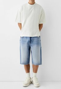 Man klädd i en vit skjorta med kort ärm, ljusblå denimshorts som går till knäna, vita ankelsockor och off-white sneakers, stående mot en vit bakgrund.